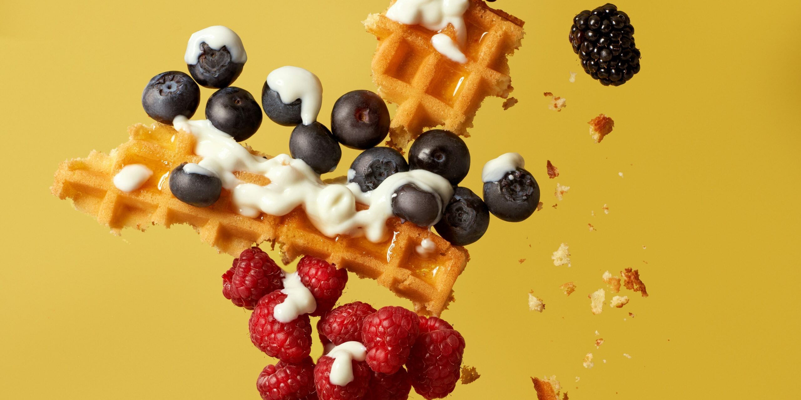 Waffle pieces with blueberries, raspberries, and blackberries topped with white yogurt, scattered against a yellow background.