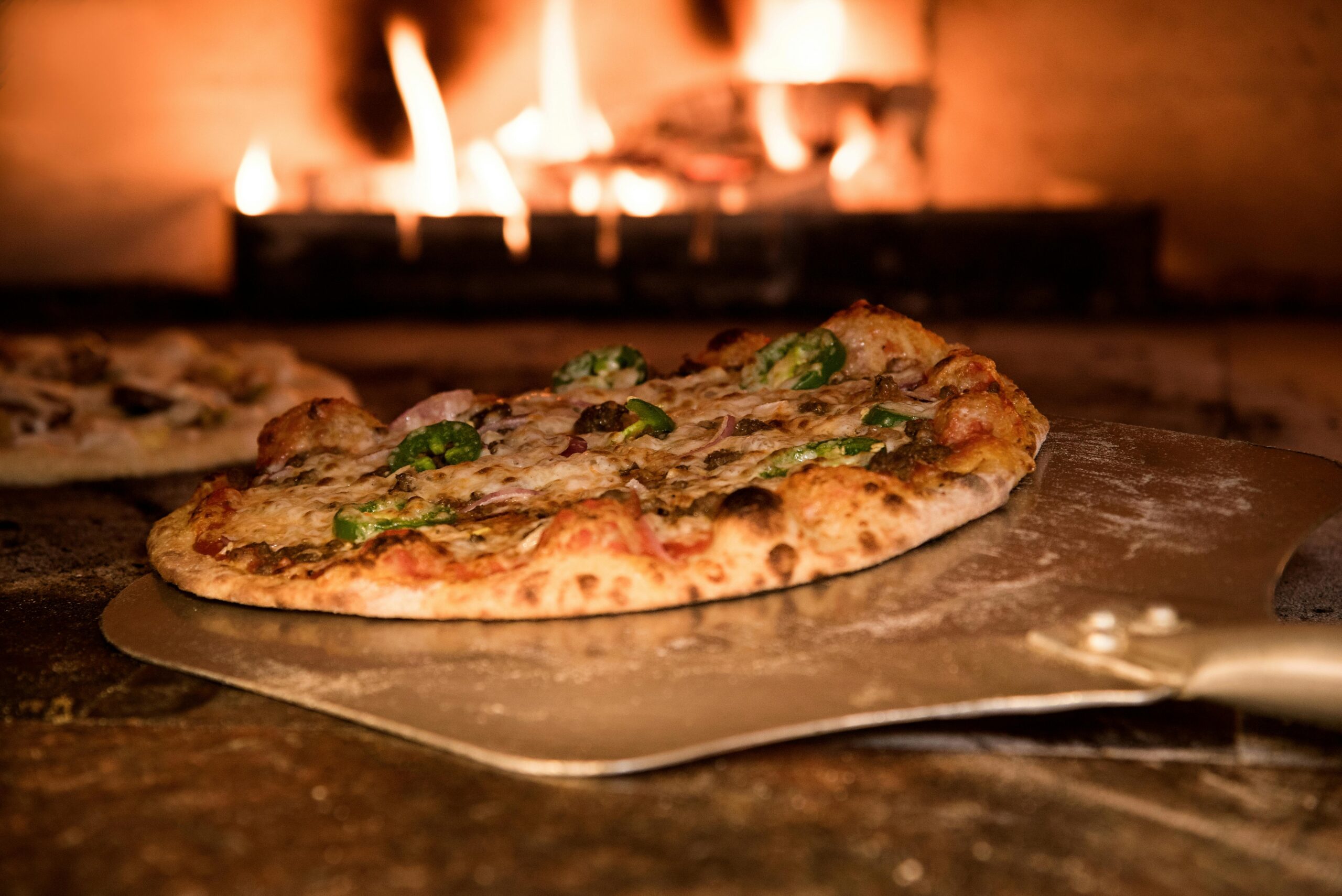 A pizza topped with various ingredients is being taken out of a wood-fired oven using a pizza peel.