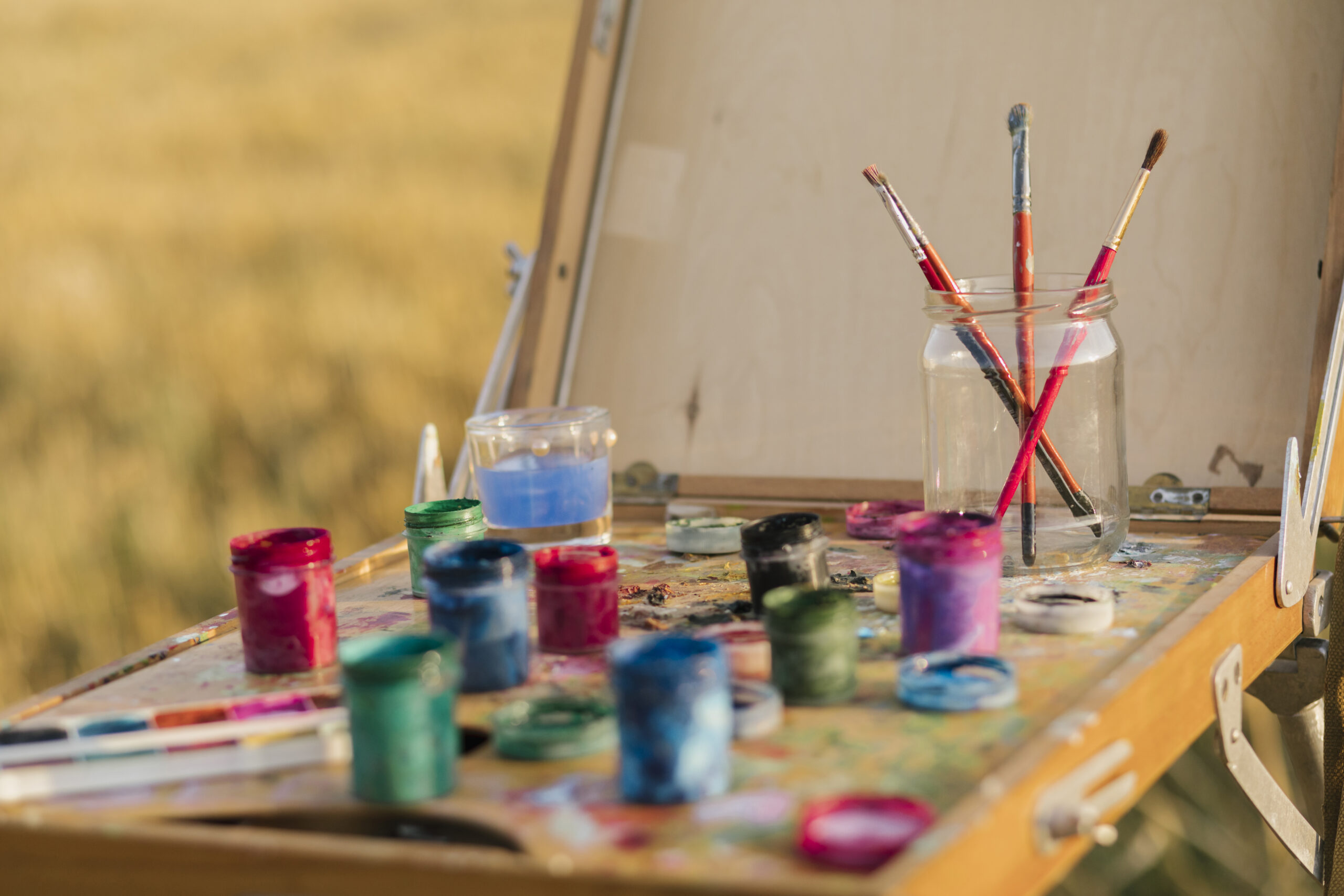 Close-up of a painter's easel with an assortment of colorful paint jars and brushes in a jar, set outdoors against a blurred background of a field.
